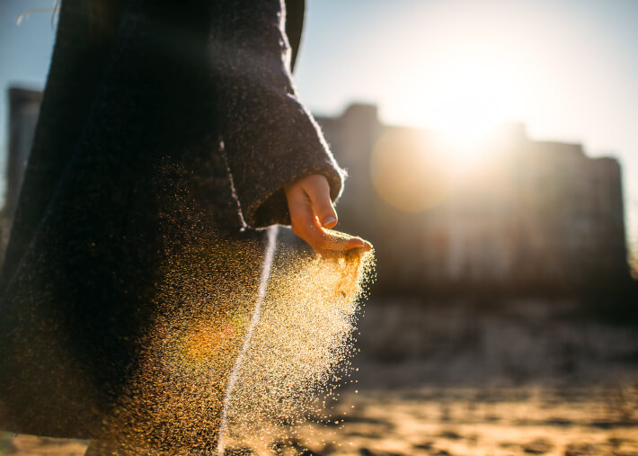 A person is walking through an arid dessert while holding a hand out that is filled with sand and slowly letting it fall between their fingers.
