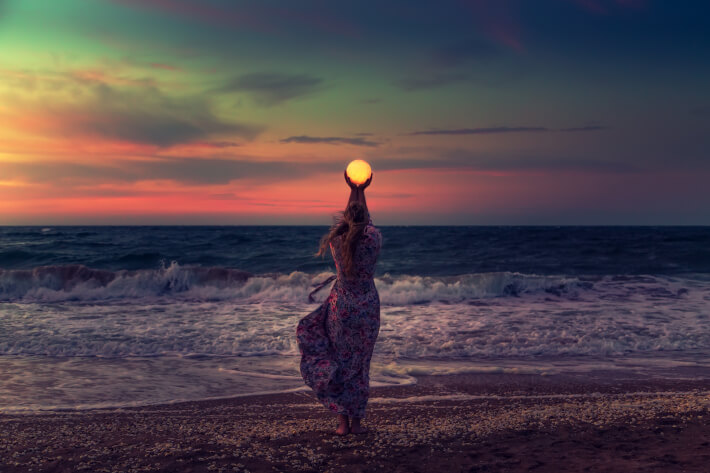 A woman in a flowing dress, on the coast of a large body of water is holding a glowing orb up to the sky.